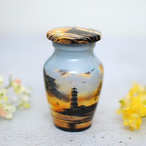 May include: A small decorative urn featuring a lighthouse scene. The urn has a light blue body with a sunset design featuring a lighthouse, water, and birds. The lid is a marbled brown and gold.