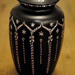 May include: A black urn with a silver lid and intricate silver floral designs. The urn has a rounded body and a slightly flared top.