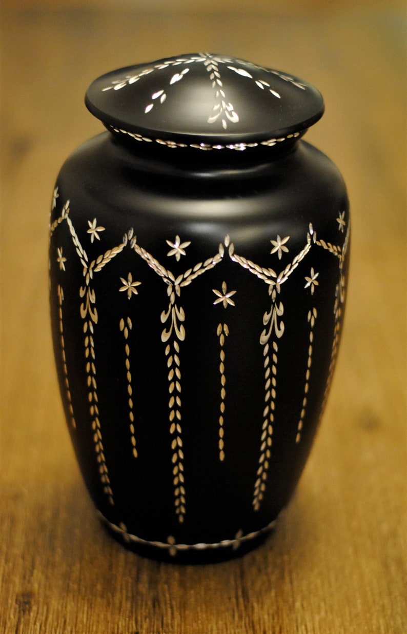 May include: A black urn with a silver lid and intricate silver floral designs. The urn has a rounded body and a slightly flared top.