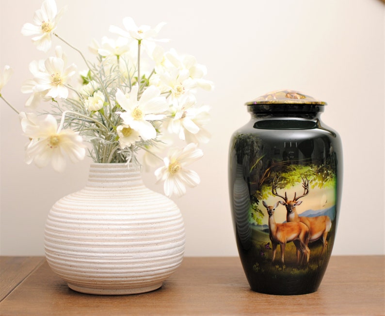 K&ouml;nnte beinhalten: Eine schwarze und gr&uuml;ne Metallurne mit einer Hirschszene. Die Urne hat einen goldenen Deckel und steht auf einer Holzoberfl&auml;che. Eine wei&szlig;e Vase mit wei&szlig;en Blumen steht im Hintergrund.
