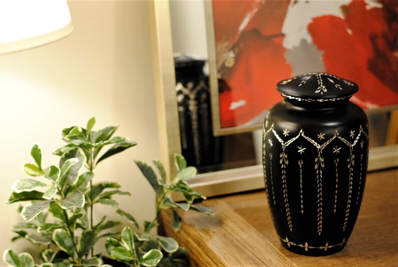 May include: Black urn with a silver design. The urn has a lid and is decorated with a geometric pattern.