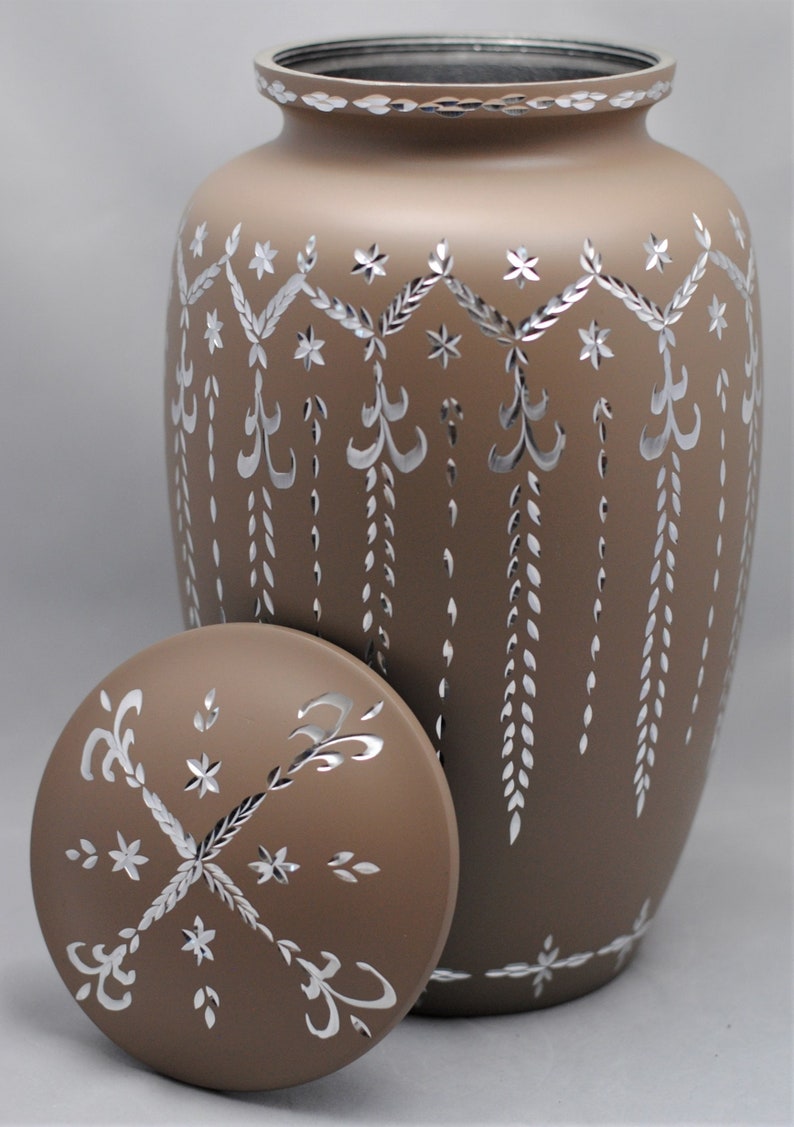 May include: A brown ceramic urn with a silver-colored design. The urn has a lid with a similar design. The urn is decorated with a repeating pattern of leaves and stars.