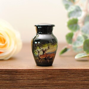 K&ouml;nnte beinhalten: Eine kleine schwarze Urne mit einem gemalten Bild von zwei Hirschen in einer Waldlandschaft. Die Urne steht auf einer Holzoberfl&auml;che mit einer wei&szlig;en Rose und gr&uuml;nem Laub im Hintergrund.