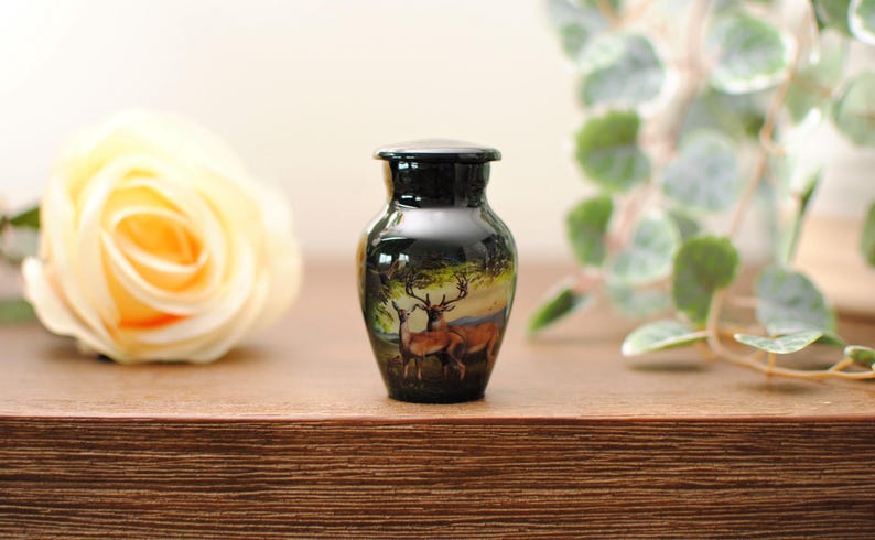 K&ouml;nnte beinhalten: Eine kleine schwarze Urne mit einem gemalten Bild von zwei Hirschen in einer Waldlandschaft. Die Urne steht auf einer Holzoberfl&auml;che mit einer wei&szlig;en Rose und gr&uuml;nem Laub im Hintergrund.