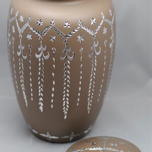 May include: A brown ceramic urn with a silver design. The urn has a lid with a similar design. The urn is decorated with a repeating pattern of stars, leaves, and swirls.