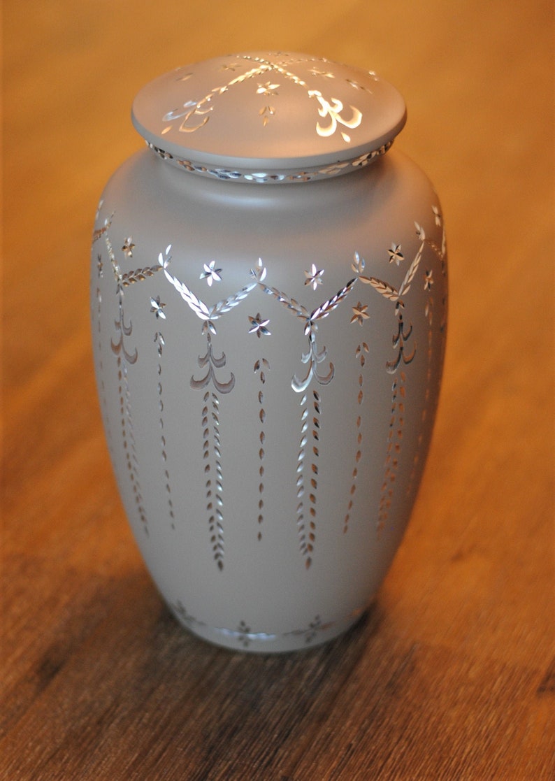 May include: A light beige ceramic urn with a silver lid and intricate silver floral designs. The urn has a smooth, matte finish.