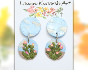May include: Handmade earrings featuring a blue sky design with white clouds and a cactus scene. The earrings are round with a silver hook. The text "Leann Kucerik Art" and a website address are printed on the card.