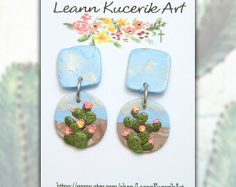 May include: Handmade polymer clay earrings featuring a desert cactus design. The earrings have square blue tops with a cloud design, and round bottoms with a cactus, pink flowers, and a desert landscape. The card reads "Leann Kucerik Art".