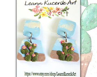 May include: A pair of handmade earrings featuring a desert cactus design. The earrings have a light blue background with a brown base, and a green cactus with pink flowers. The card reads "Leann Kucerik Art" and the Etsy shop URL.