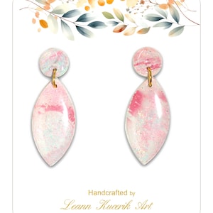 May include: A pair of handcrafted earrings with a pink and white marbled design. The earrings have a teardrop shape with a small circular top, connected by a gold-colored loop. The card reads "Handcrafted by Leann Kucerik Art."
