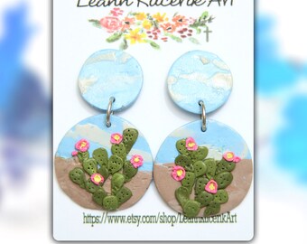 May include: Handmade polymer clay earrings featuring a desert landscape design. The earrings have a light blue sky, a brown desert base, and green cacti with pink flowers. The top portion is a light blue circle with white accents. The text "Leann Kucerik Art" is visible.