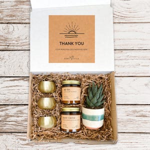 May include: A gift box filled with brown packing paper, containing three gold candle holders, two candles labeled "Red Sea" and "Black Cactus", and a small potted succulent plant. The box lid has a brown card with a sun and waves graphic and the text "Thank You, Your personalized message here, Rebel Villa".