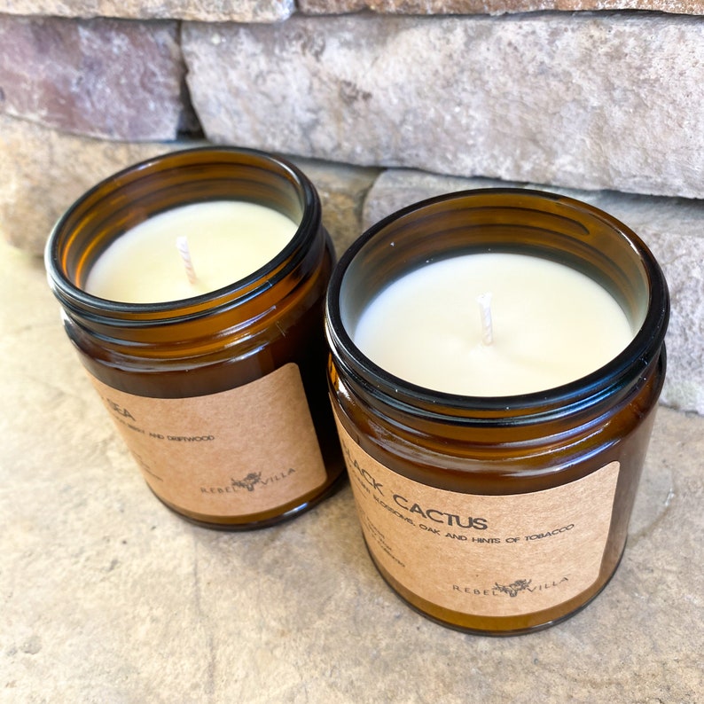 May include: Two amber glass candle jars with brown paper labels. The candle on the left is labeled "TEA" with a small image of a deer. The candle on the right is labeled "BLACK CACTUS" with a small image of a deer. The candle on the right has a white wax surface and a white wick.