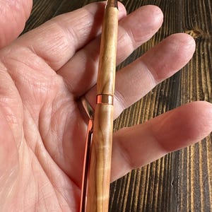 May include: A wooden pen with a copper finish. The pen has a natural wood grain pattern and a copper colored clip and tip.