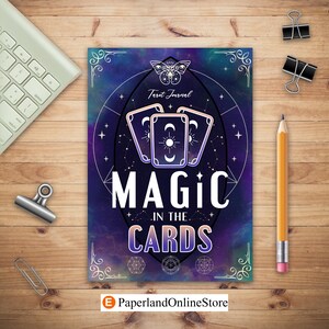 May include: A tarot journal with a celestial design. The cover features three tarot cards, a butterfly, and the words "MAGIC IN THE CARDS." The background is a gradient of blues and purples. A pencil, paper clips, and a keyboard are on a wooden surface.