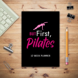 May include: A black planner with a pink silhouette of a person doing pilates. The text "BUT First, Pilates" is in pink and the text "12 WEEK PLANNER" is in white.