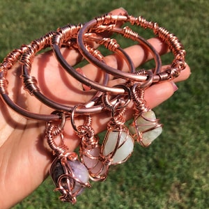 May include: Copper wire bracelets with gemstone pendants. Each bracelet features a copper wire band with wrapped accents. The pendants are various colors, including purple, pink, and green, and are wire-wrapped to the bracelets.