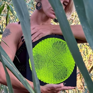 Op de afbeelding: Een zwart vierkant string art-stuk met een helder neon groen cirkelvormig ontwerp. Het kunstwerk wordt door een persoon vastgehouden, met een achtergrond die door bladeren is vervaagd. Moderne, geometrische wanddecoratie.