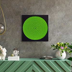 Könnte beinhalten: Ein grünes Mandala-Kunstwerk auf einer schwarzen quadratischen Leinwand. Das Kunstwerk wird auf einem grünen Schrank mit dekorativen Elementen ausgestellt, darunter eine weiße Katzenfigur, eine Uhr und Vasen mit Blumen. Über dem Ganzen hängt eine goldene Leuchte.