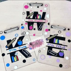 May include: Three clear acrylic clipboards with black letters, colorful accents, and teacher names. The clipboards feature a pencil, apple, and books. The names on the clipboards are "Mrs. Wiggleton", "Mrs. King", and "Mrs. Elliot".