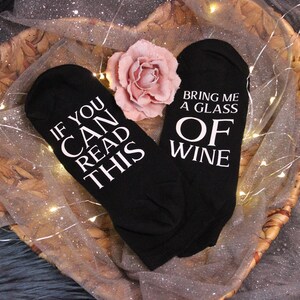 Könnte beinhalten: Schwarze Socken mit weißem Text: „IF YOU CAN READ THIS“ und „BRING ME A GLASS OF WINE“. Die Socken liegen in einem Weidenkorb, verziert mit einer rosa Rose und Lichterketten.