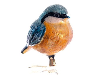 Puede incluir: Adorno decorativo de pájaro con cabeza azul grisáceo, detalles en blanco y negro y cuerpo naranja dorado. El pájaro tiene una pequeña campana en la base y está sobre un fondo blanco. Una encantadora decoración navideña.