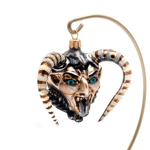 Puede incluir: Un adorno de Navidad de vidrio con forma de cabeza de diablo con cuernos rayados en dorado y blanco. El adorno tiene ojos azules y la lengua fuera. El adorno está colgado de un soporte de metal dorado.