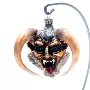 Puede incluir: Un adorno de Navidad de vidrio con forma de cabeza de demonio con cuernos, ojos azules y lengua roja. El adorno está colgado de un gancho plateado.