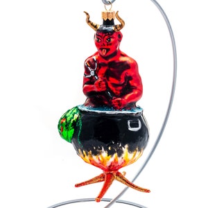 Puede incluir: Un adorno de Navidad de vidrio rojo que representa a un diablo revolviendo un caldero con un tenedor. El adorno está en un soporte plateado con una base redonda.