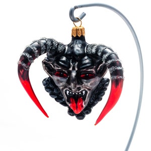 Puede incluir: Un adorno de Navidad de vidrio negro y rojo con forma de cabeza de diablo con cuernos. El adorno tiene ojos rojos y una lengua roja que sobresale.