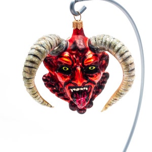 Puede incluir: Un adorno navideño rojo y negro con cara de diablo, cuernos grandes y curvados y ojos amarillos. El adorno tiene una expresión amenazante con la lengua fuera. Está suspendido de un gancho de metal plateado.