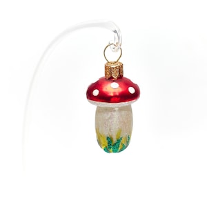Puede incluir: Un adorno de Navidad de vidrio rojo y blanco con forma de hongo con detalles verdes y amarillos. El adorno tiene un lazo dorado para colgar.