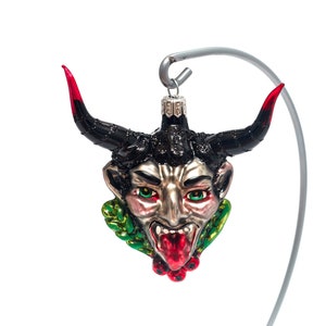 Puede incluir: Un adorno navideño con temática de diablo con una cara plateada, ojos verdes y cuernos con puntas rojas. El adorno tiene una melena negra y una lengua roja. Está suspendido de un gancho plateado.