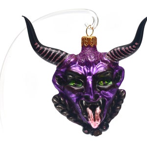 Puede incluir: Adorno de Navidad de vidrio morado con forma de cabeza de diablo con cuernos negros y una lengua rosa. El adorno tiene detalles de purpurina y detalles negros alrededor de la cara.