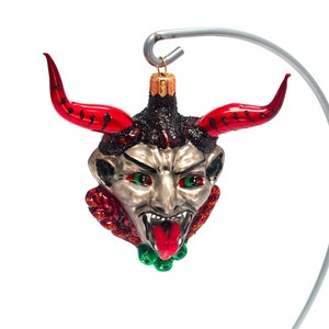 Puede incluir: Un adorno de Navidad de vidrio rojo y plateado con forma de cabeza de diablo con ojos verdes y lengua roja.