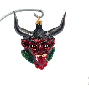 Puede incluir: Adorno de Navidad de vidrio rojo y negro con forma de cabeza de diablo con cuernos, que lleva una corona de acebo verde y bayas rojas.