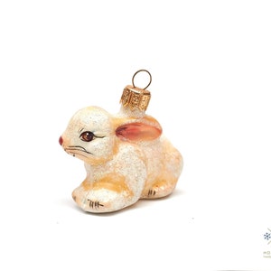 以下が含まれることがあります： 金色のトップが付いた、白いガラス製のウサギの形をしたクリスマスオーナメント。ウサギはピンクの耳と茶色の目を持っています。