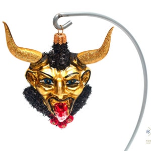 Puede incluir: Un adorno navideño dorado y negro que representa la cara de un diablo con cuernos, barba negra y lengua roja. El adorno está colgado de un gancho plateado.