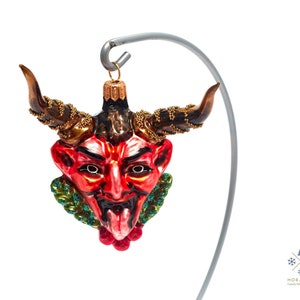Puede incluir: Adorno de Navidad de vidrio rojo y negro con forma de cara de diablo con cuernos, detalles dorados y follaje verde y rojo.