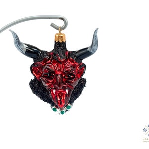 Puede incluir: Adorno de Navidad de vidrio rojo con forma de cabeza de diablo con cuernos negros y un collar de joyas verdes.