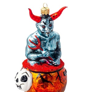 Puede incluir: Un adorno de Navidad de vidrio que representa un diablo azul con cuernos rojos y un tridente sentado en un caldero con una calavera y llamas. El adorno tiene una estrella dorada en la parte superior y una base dorada.