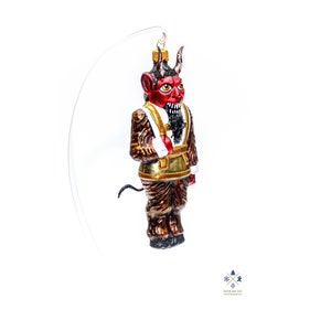 Puede incluir: Adorno navideño rojo y dorado que representa un diablo con cuernos, barba y ribete de piel blanca. El adorno está colgado de una cuerda transparente.