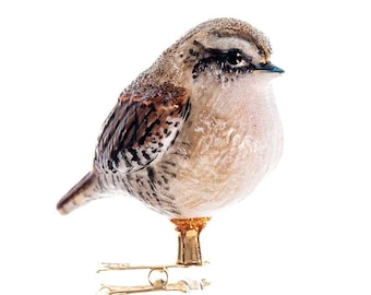 Puede incluir: Un adorno de vidrio de un pájaro marrón y blanco con un soporte dorado. El pájaro está posado en el soporte con la cabeza girada hacia un lado.