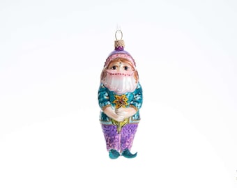 May include: A glass ornament shaped like a gnome wearing a purple hat and pants, a teal jacket with white leaf patterns, and a white beard. The gnome holds a yellow flower and has a small loop at the top for hanging on a tree.