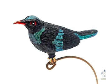 Puede incluir: Adorno de pájaro negro con detalles de purpurina turquesa en la cabeza, alas y cola. El pájaro tiene un pico rojo y ojos rojos. Está posado sobre un soporte de metal dorado, ideal para la decoración navideña.