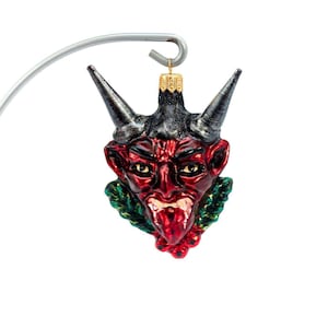 Puede incluir: Un adorno navideño con cara de diablo rojo y negro con cuernos, una expresión de mueca y un cuello verde y rojo. El adorno está suspendido de un gancho de metal plateado. Una decoración festiva.
