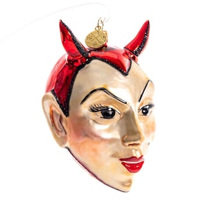 Puede incluir: Adorno decorativo con forma de cabeza de diablo. El adorno presenta cuernos rojos, pelo rojo y una cara clara con labios rojos. Se suspende mediante un lazo dorado.