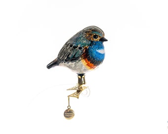 Puede incluir: Adorno de pájaro de cristal detallado con plumaje azul, naranja y gris. El pájaro está posado sobre un clip dorado con un pequeño dije redondo que dice "HOBBY CHERISH". El adorno está sobre un fondo blanco.