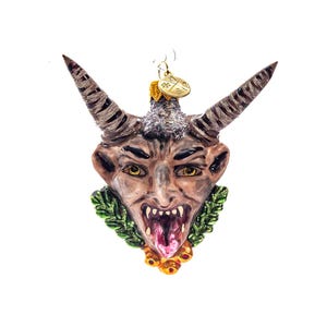 Puede incluir: Un adorno de Navidad de vidrio que representa la cabeza de un diablo verde y marrón con una lengua larga y cuernos. El adorno está decorado con hojas verdes y bayas rojas.