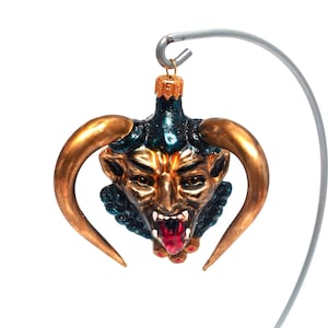 Puede incluir: Un adorno de Navidad de vidrio con forma de cabeza de diablo con cuernos dorados, una cara verde y azul y una lengua roja.